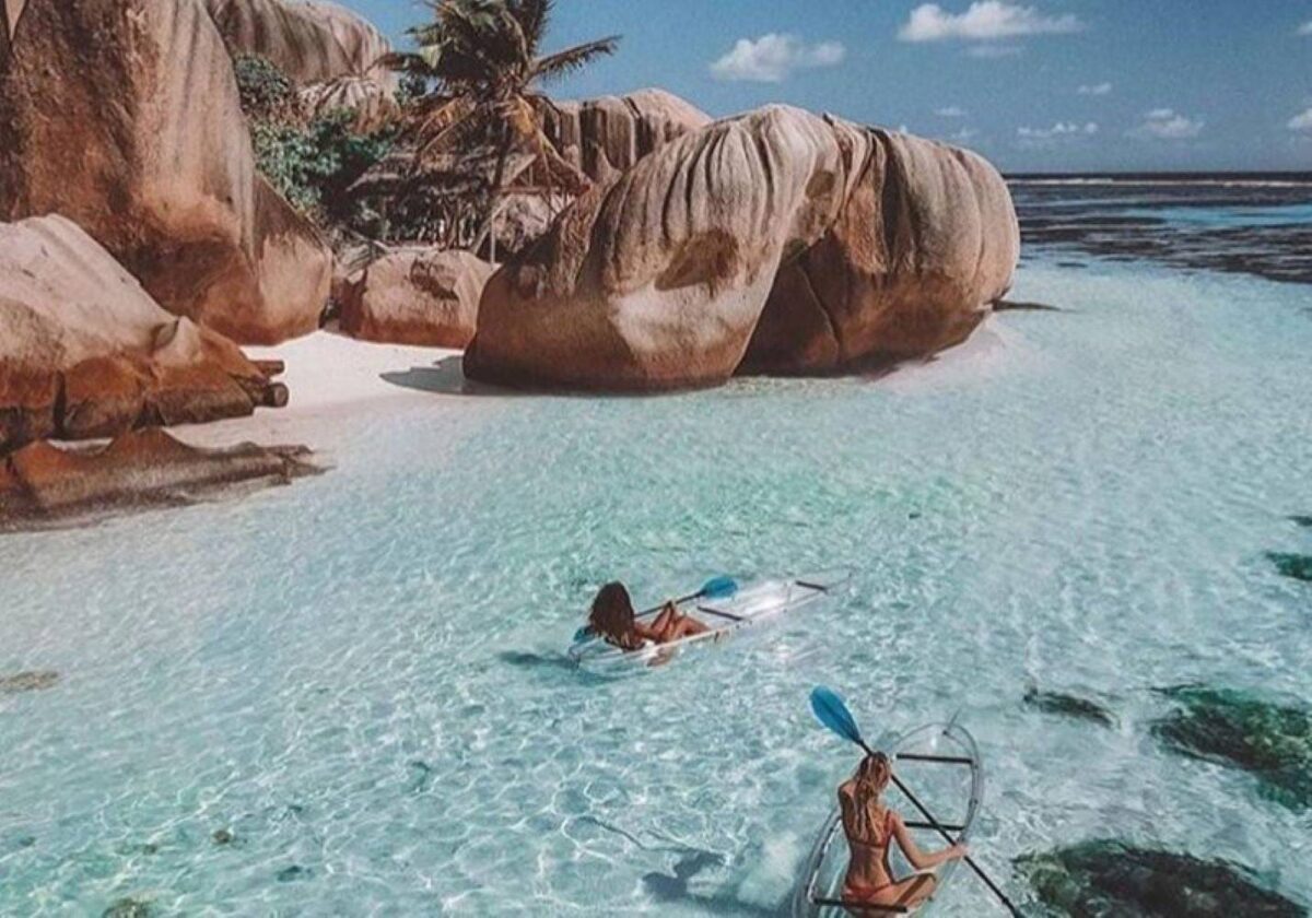 Couple paddling in a transparent kayak over clear turquoise waters off La Digue, with granite rocks in the background