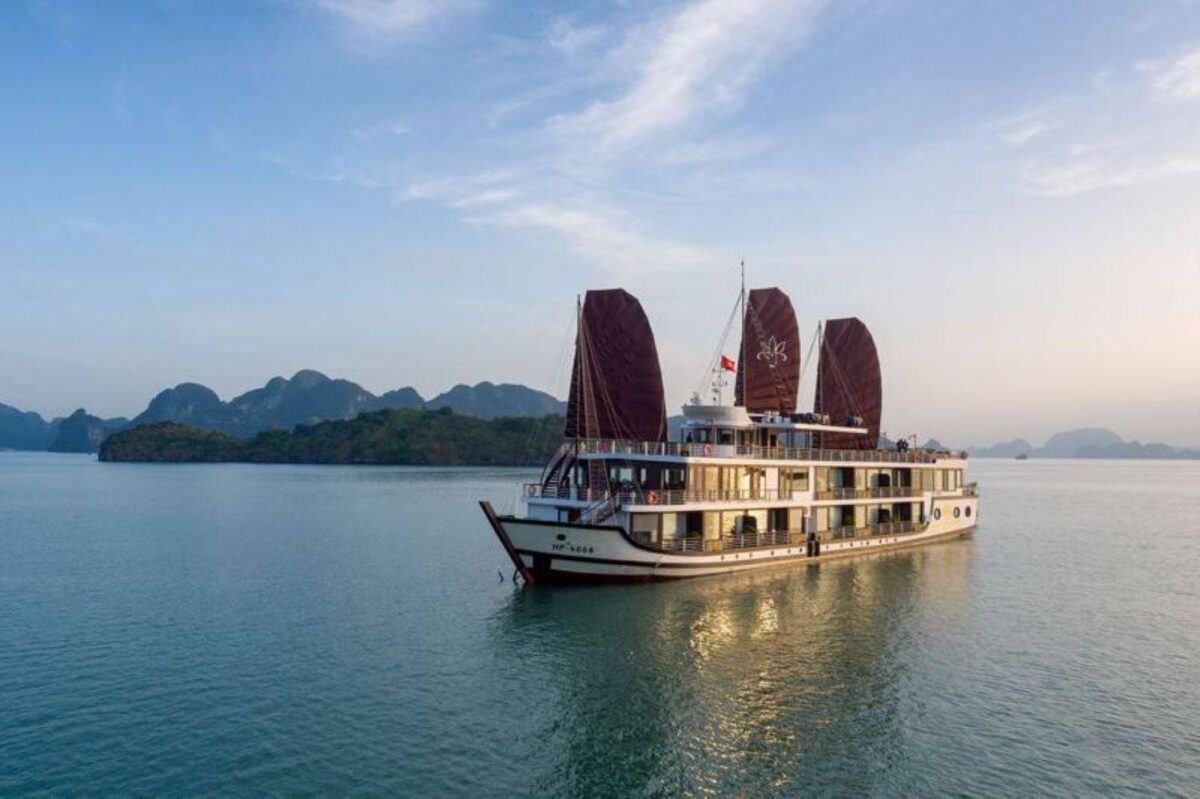 Cruise ship sailing between steep limestone karst islands in Ha Long Bay under a hazy sky with calm emerald water