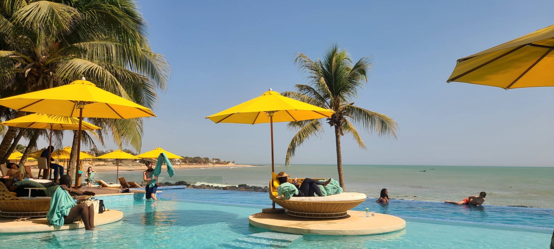 Family walking along a quiet sandy beach in Somone, Senegal, with palm trees, calm waves and a relaxed seaside hotel in the background