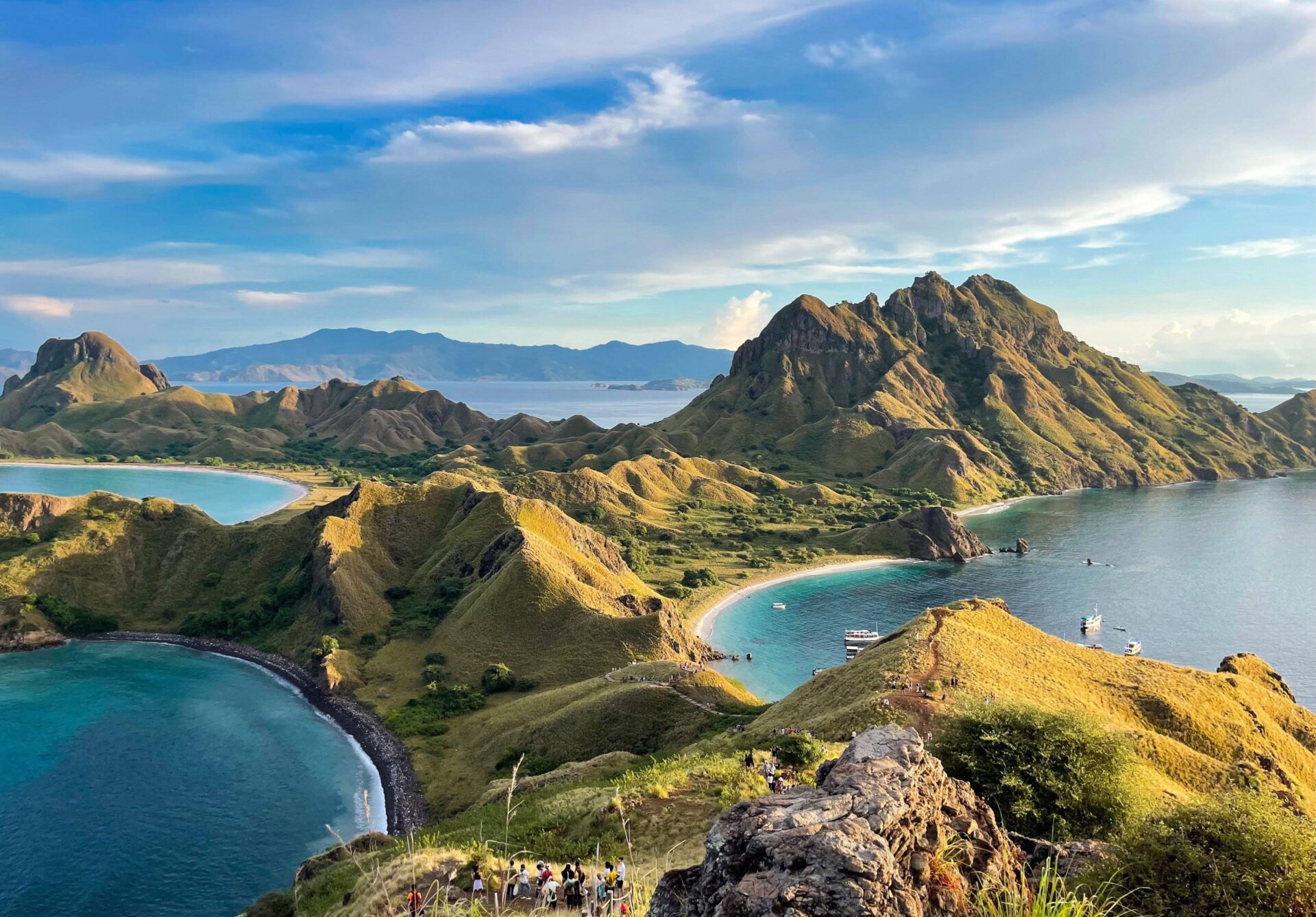Panoramic view of a Balinese coastal temple on a rocky outcrop at sunset with waves crashing against the cliffs and lush greenery in the background