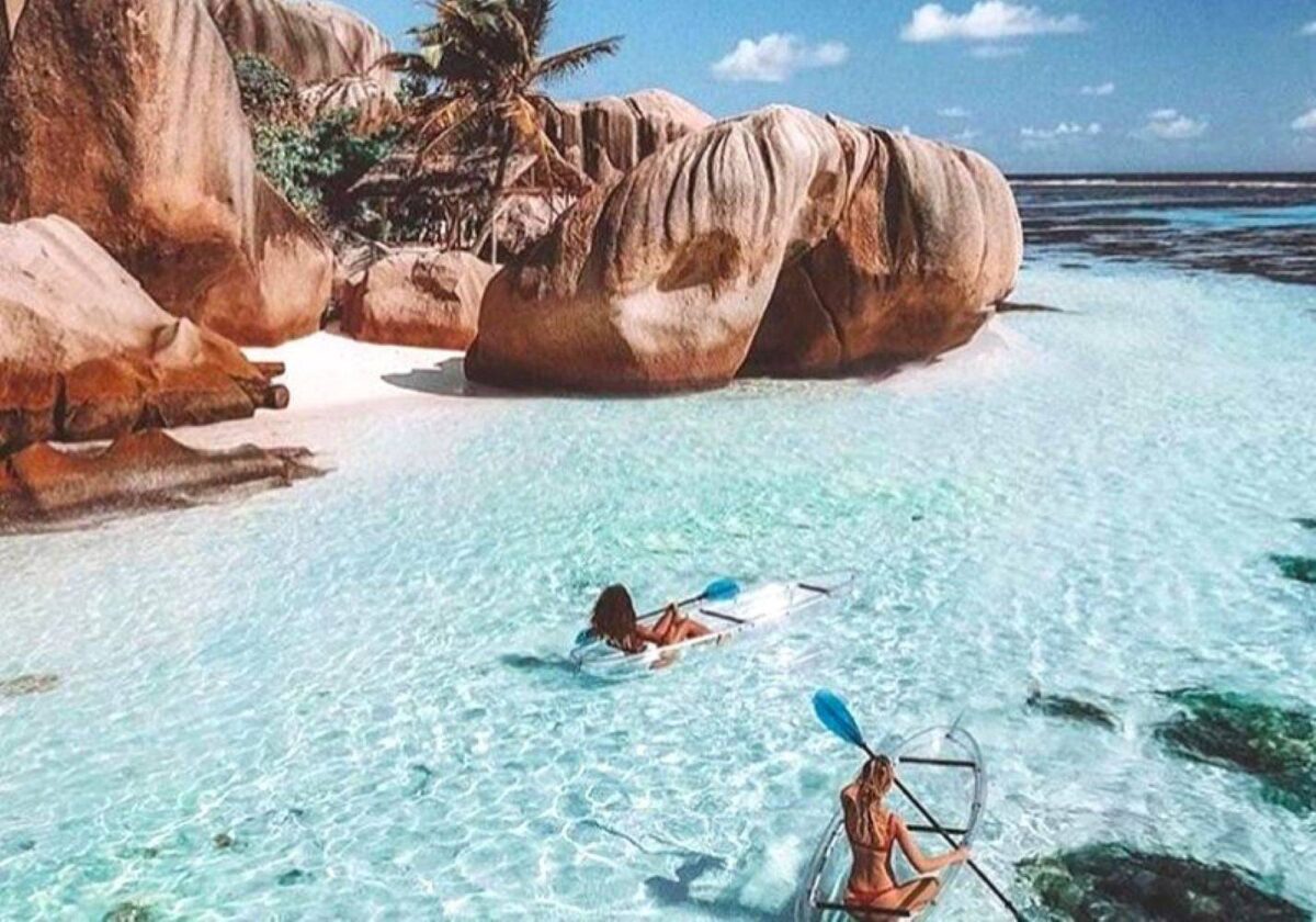 Person paddling a transparent kayak over clear turquoise water above coral near La Digue