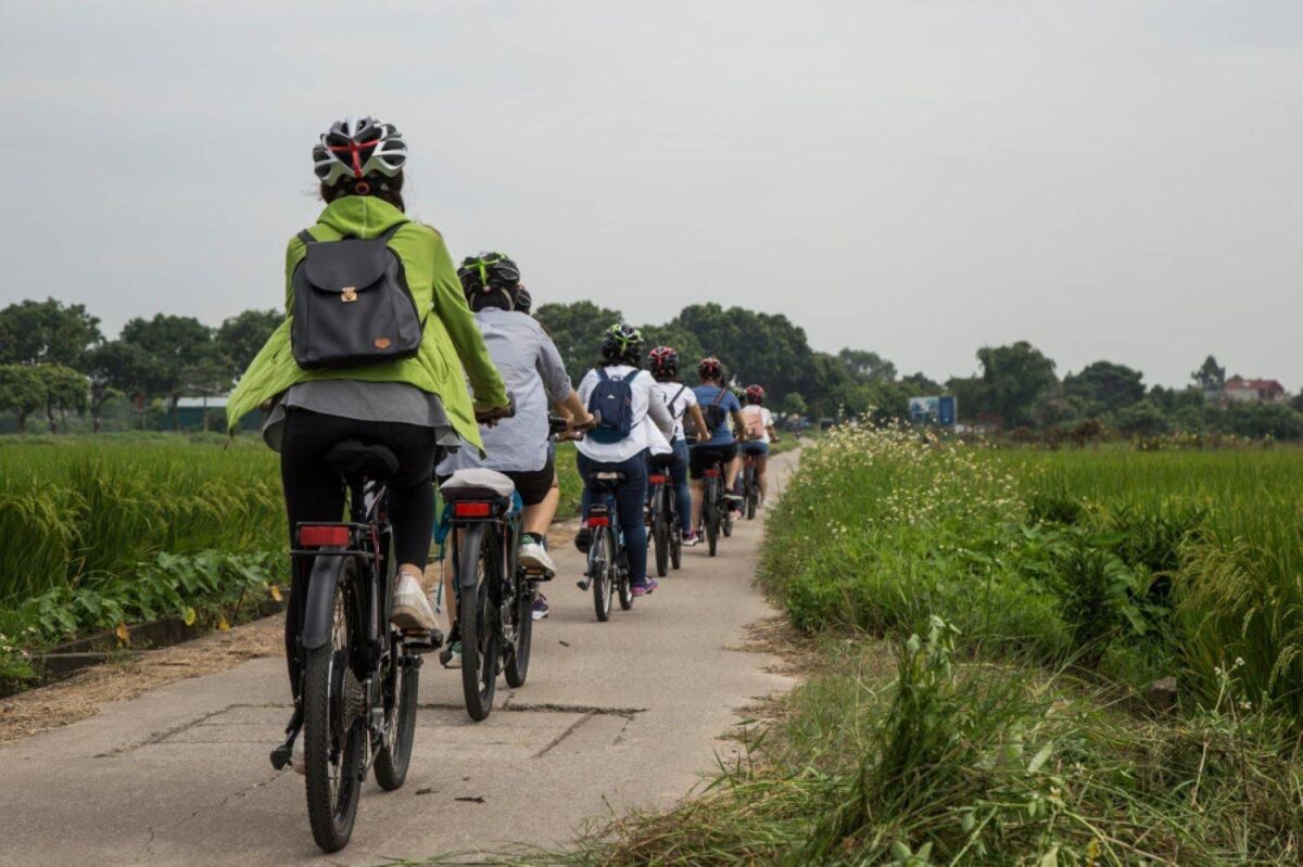 Two cyclists riding along a rural path near Hanoi past green rice paddies and small village houses under an overcast sky